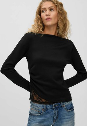 Femme aux cheveux blonds bouclés portant un haut noir à manches longues avec des détails en dentelle et un jean bleu, regardant sur le côté, les mains dans le dos.