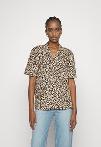 Camisa de manga curta com padrão de leopardo em tons de bege e castanho, apresentando um colarinho com recorte e um corte descontraído. Combinada com jeans de tom azul claro.