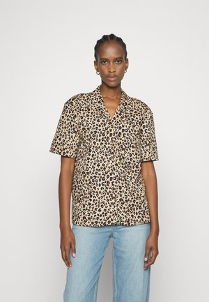 Camisa de manga curta com padrão de leopardo em tons de bege e castanho, apresentando um colarinho com recorte e um corte descontraído. Combinada com jeans de tom azul claro.