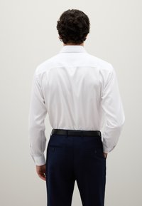 Witte blouse met lange mouwen en een klassieke kraag, gecombineerd met marineblauwe geruite broek, voorzien van een zwarte riem en subtiele achterdetails.