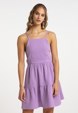 Frau mit langen Haaren, die große bunte Ohrringe und ein ärmelloses lilafarbenes Kleid mit dünnen Schulterträgern vor weißem Hintergrund trägt.