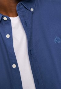 Niebieska koszula zapinana na guziki z kołnierzykiem, ozdobiona białymi guzikami i jasnoniebieskim logo. Pod spodem widoczna jest gładka biała koszulka z okrągłym dekoltem.