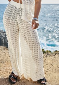 Pantalones de pierna ancha blancos de punto con un patrón en forma de chevron. Combinados con sandalias negras con hebillas y tachuelas. El fondo muestra el océano.
