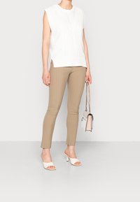 Maglione senza maniche a treccia bianco sopra una maglietta bianca, abbinato a pantaloni beige a righe sottili e mule open-toe bianchi con tacco, mentre tiene una borsa.