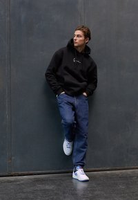 Svart hettegenser med frontend lomme, blå jeans og hvite joggesko med blå detaljer, stående mot en grå vegg.