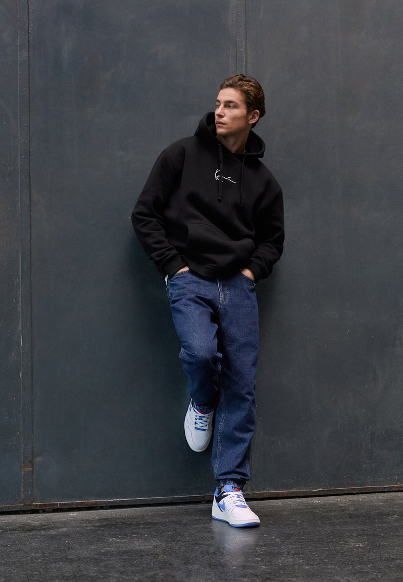 Svart hettegenser med frontend lomme, blå jeans og hvite joggesko med blå detaljer, stående mot en grå vegg.