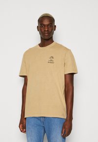 Beige cotton t-shirt with short sleeves, round neck, and graphic print. Paired with blue jeans. Soft texture and relaxed fit.