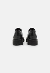 Chaussures en cuir noir avec une finition lisse, bout arrondi et semelles en caoutchouc noir chunky présentant un motif de semelle, vues de l'arrière.