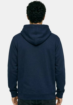 Homme aux cheveux noirs bouclés portant un sweat à capuche bleu marine uni, vu de dos sur un fond blanc.