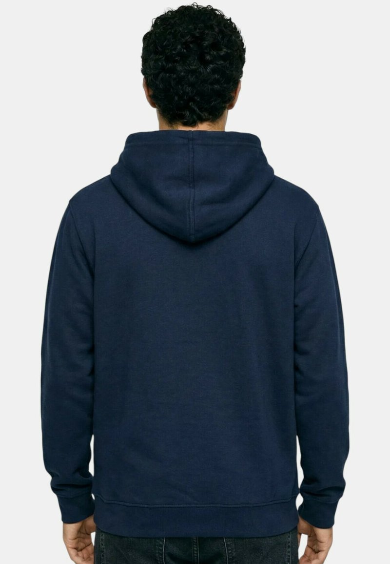 Homme aux cheveux noirs bouclés portant un sweat à capuche bleu marine uni, vu de dos sur un fond blanc.