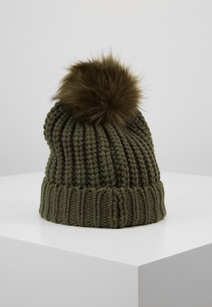 Bonnet tricoté vert foncé avec de grosses côtes et un grand pompon marron duveteux sur le dessus, posé sur une surface blanche.