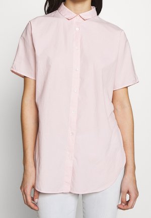 Femme portant une chemise rose clair à manches courtes boutonnée avec un col classique et un pantalon blanc.