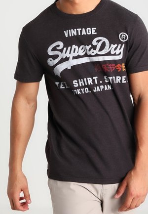 Mężczyzna w czarnej koszulce z białym i czerwonym napisem "SuperDry Vintage T-Shirt Store Tokyo, Japan", w jasnobeżowych spodniach.