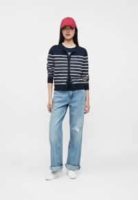 Μπλε ριγέ κάρντιγκ με κουμπιά, φορεμένο πάνω από μπλε μπλουζάκι και συνδυασμένο με ανοιχτό μπλε τζιν με σκισίματα και λευκά αθλητικά παπούτσια, ολοκληρωμένο με κόκκινο καπέλο.