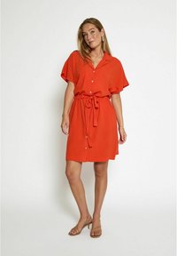 Red short-sleeve dress with a collar, buttons down the front, tie waist, and soft texture. Worn with strappy sandals.