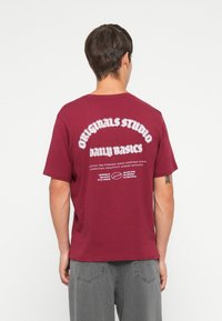 Burgunderfarget bomulls t-skjorte med hvit grafisk tekst på baksiden, korte ermer og løs passform. Stoffet ser mykt og glatt ut.