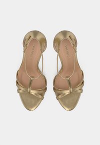 Pari avokatoja metalike të arta me pësoh të hapura, me shirita rreth kyçit dhe detaje të kryqëzuara, të parë nga lart mbi një sfond të bardhë.