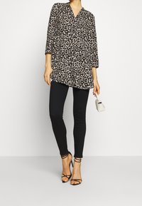 Tunique à imprimé léopard avec manches trois-quarts, associée à des leggings noirs et des talons noirs à brides ; tenant un petit sac à main blanc.