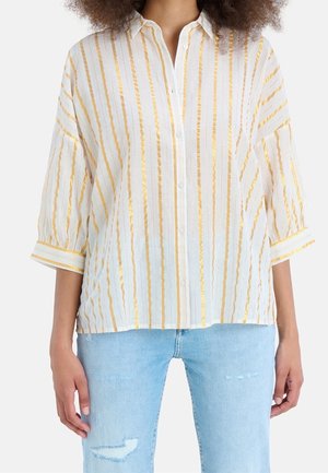 Mujer con una camisa blanca abotonada con rayas doradas verticales y vaqueros desgastados azul claro, mostrada desde el cuello hasta las caderas.