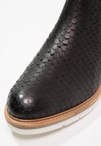 Botas de cuero negro con un patrón texturizado en forma de escamas de pez; punta redondeada; realzadas por una suela marrón y blanca, creando un diseño distintivo.