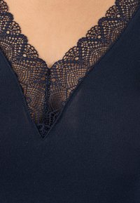 Donkerblauwe tanktop met een diepe V-hals en intrigerende kantafwerking, gemaakt van een zachte, rekbare stof. De kant heeft bloemmotieven langs de rand.