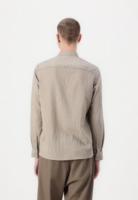Beige hosszú ujjú csíkos ing, gallérral, gombos mandzsettával és textúrált anyagból, laza barna nadrággal viselve, hátulról nézve.