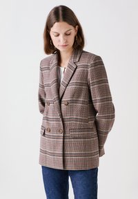 Bruine geruite blazer met dubbele knoopsluiting, geknipte revers, voorzakken en bruine knopen. Draagt over een witte top en blauwe spijkerbroek.