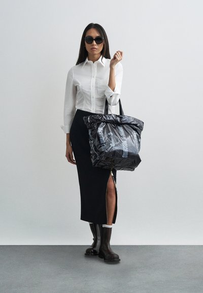 Μαύρη τσάντα tote με γυαλιστερή, τσαλακωμένη υφή και λευκό λογότυπο. Το μοντέλο φοράει λευκό πουκάμισο με κουμπιά, μαύρη midi φούστα και καφέ μποτάκια μέχρι τον αστράγαλο.