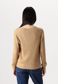 Pull en tricot texturé beige avec un col rond et des poignets côtelés, vu de dos, associé à un jean bleu foncé.