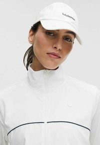 Valge tõmblukkudega jakk kõrge kraega, tumesiniste aksentjoontega ja logo nööbiga. Täiendatud valge mütsiga, millel on must brändimine.