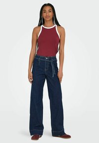 Top sin mangas de canalé marrón con ribete blanco, combinado con jeans oscuros de pierna ancha y talle alto, que cuentan con un cinturón anudado y una textura clásica de denim.