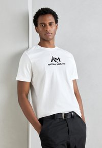 T-shirt en coton blanc avec un logo graphique noir "ANTHONY MORATO" sur la poitrine, associé à un pantalon sombre et une ceinture noire.