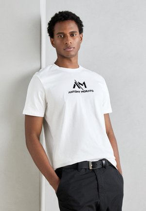 T-shirt en coton blanc avec un logo graphique noir "ANTHONY MORATO" sur la poitrine, associé à un pantalon sombre et une ceinture noire.