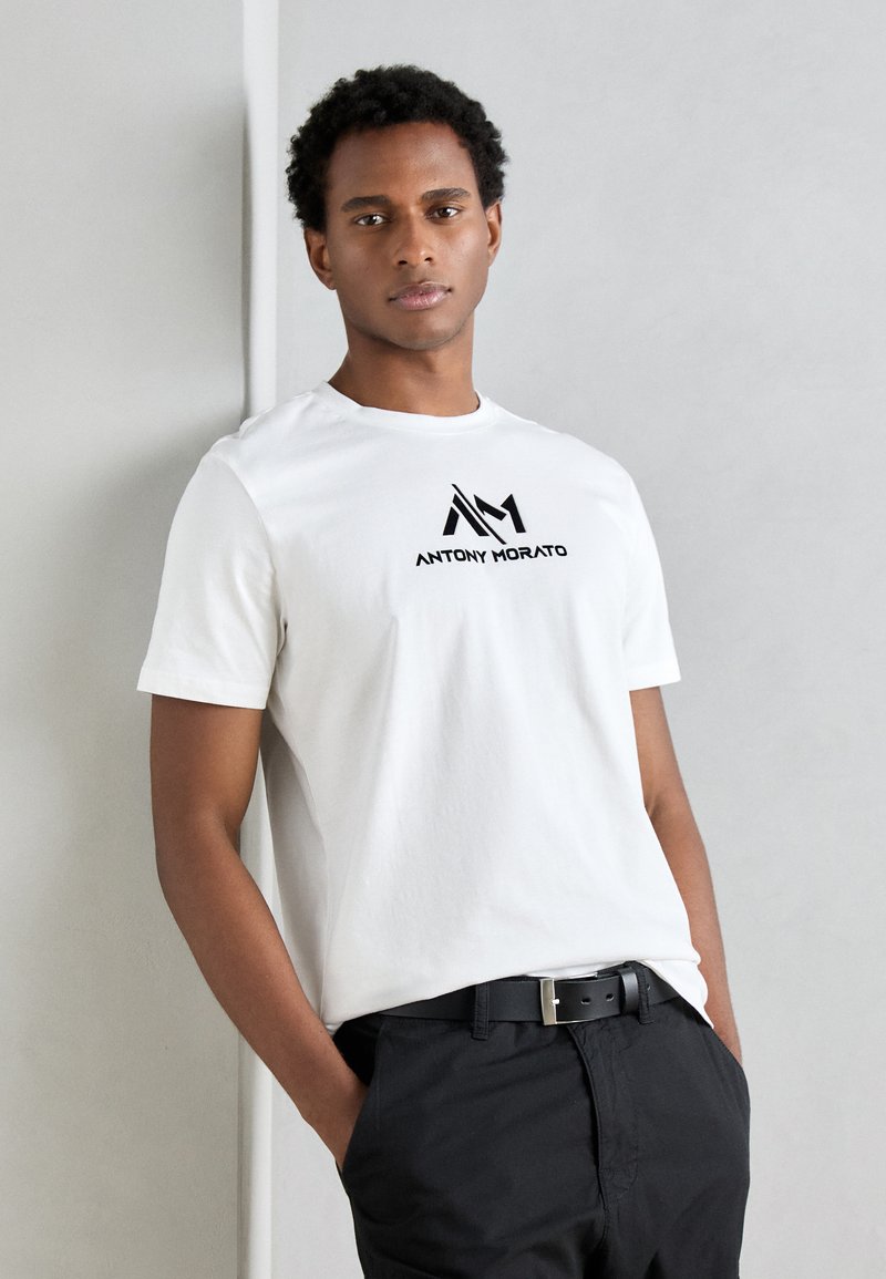 T-shirt en coton blanc avec un logo graphique noir "ANTHONY MORATO" sur la poitrine, associé à un pantalon sombre et une ceinture noire.