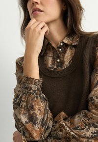 Pruunist kootud vest on kihistatuna lillemustrilise pluusi kohal, millel on puhvblööd ja elastsed varrukad. Pluusil on soojad toonid ja pärl-okkidega nööbid.