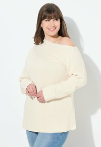 Ecrufarbener, gerippter Pullover mit asymmetrischem Ausschnitt und langen Ärmeln, kombiniert mit hellblauen Jeans. Weiche Strickstruktur sichtbar.