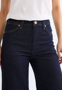 Person wearing high-waisted dark blue denim jeans with copper buttons and yellow stitching, paired with a white tucked-in top.