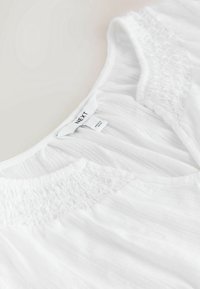Blouse blanche texturée avec des détails smockés sur les épaules et une étiquette de marque visible cousue à l'intérieur de l'encolure.
