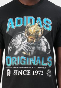 Fekete pamut póló, amelyen egy kéz tart egy trófeát, ami egy földgömbre van ráhelyezve. Kék és fehér szöveg: "ADIDAS ORIGINALS SINCE 1972."