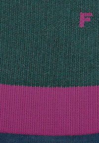 Stricksocke mit dunkelgrünem Körper, ausgestattet mit einem horizontalen pinken Band und einem blauen Band am unteren Ende. Beinhaltet ein kleines pinkes 'F'-Logo.