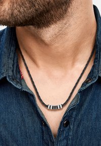 Collana in pelle nera intrecciata con accenti metallici, indossata sopra una camicia di denim, che mette in risalto il collo e l'area del collo di un uomo.