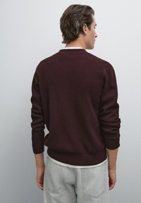 Uomo in piedi rivolto di spalle, che indossa un maglione a coste bordeaux scuro sopra una camicia bianca e pantaloni grigio chiaro, su uno sfondo grigio uniforme.