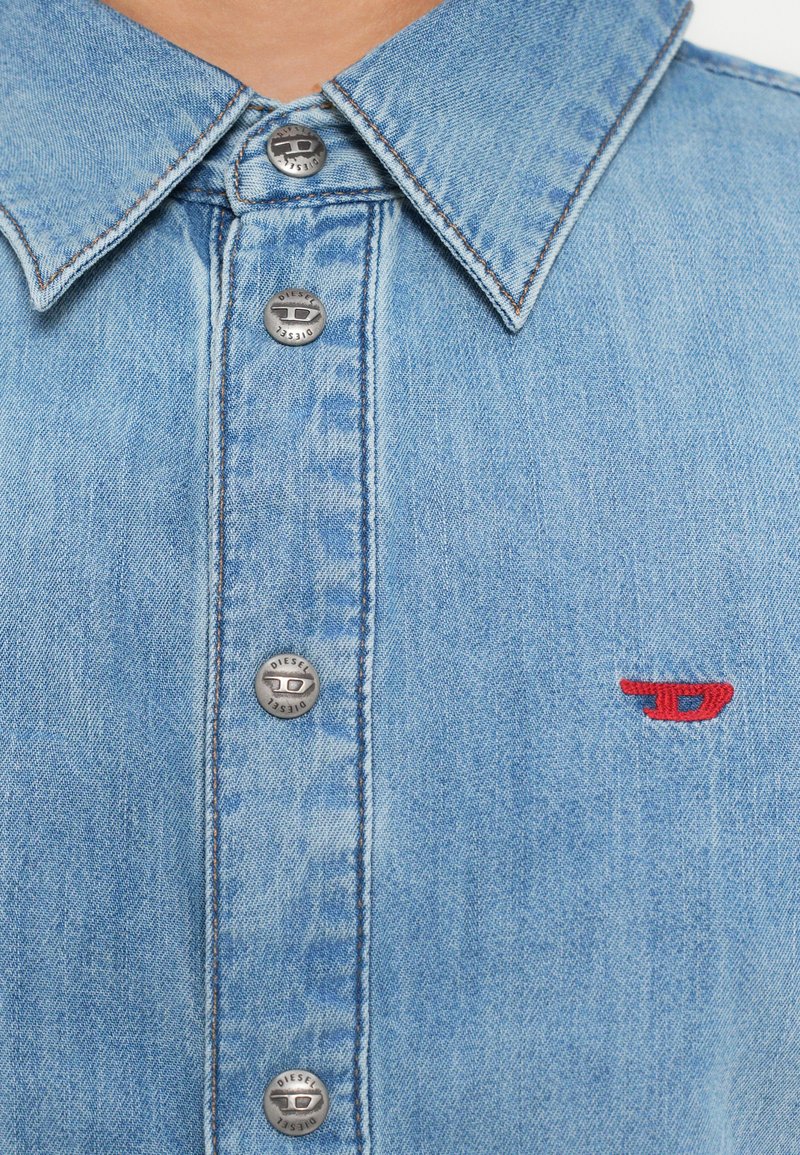 Lysblå denimskjorte med knapper, metalknapper, kontrastsyning og en rød broderet logo på venstre bryst.