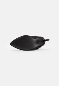 Chaussure à talon haut en daim noir avec un bout pointu et une semelle texturée, présentant un design élégant et une finition lisse sur la partie supérieure.