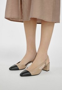 Beżowe skórzane buty slingback z czarnymi spiczastymi noskami i słupkowym obcasem. Otwarta konstrukcja z bokami oraz gładka faktura podkreślają design.