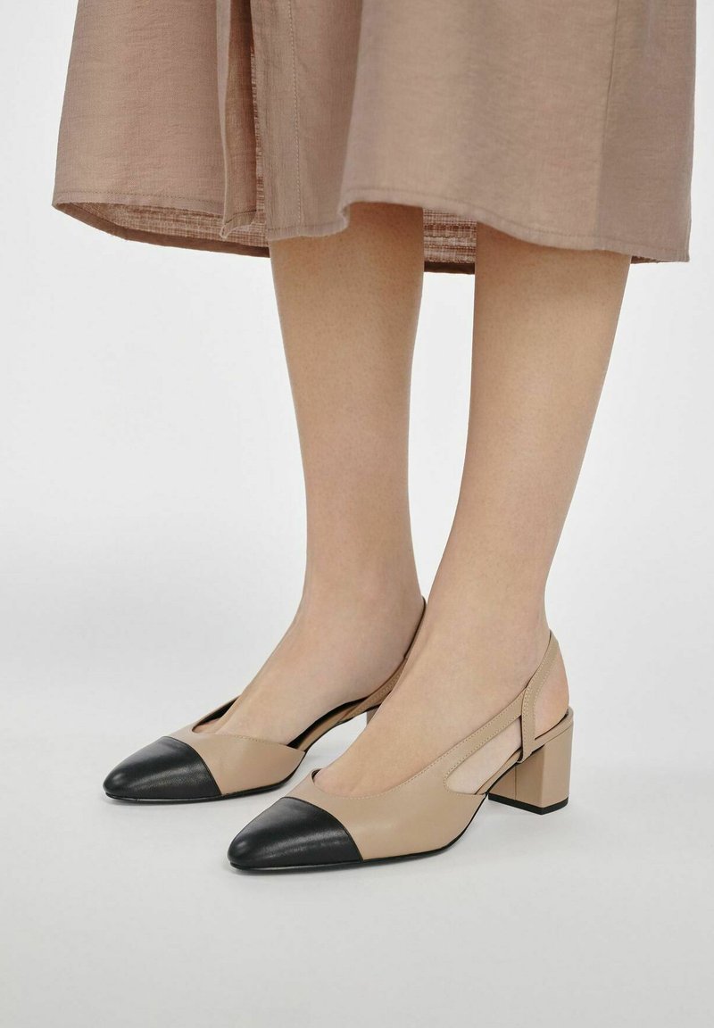Beżowe skórzane buty slingback z czarnymi spiczastymi noskami i słupkowym obcasem. Otwarta konstrukcja z bokami oraz gładka faktura podkreślają design.