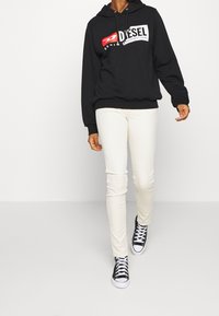 Sudadera negra con un gran logotipo "DIESEL" en blanco y rojo, combinada con pantalones blancos ajustados y zapatillas negras con suelas y cordones blancos.