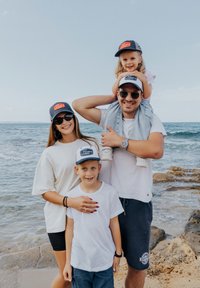 Family at the beach, wearing matching dark blue caps with orange logos. They are dressed in casual white and blue clothing, standing on rocky shore.