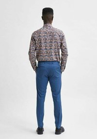 Barevná vzorovaná košile z látky s dlouhými rukávy, kombinovaná s modrými kalhotami a černými botami, pohled zezadu.