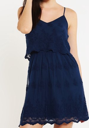 Woman wearing a sleeveless navy blue lace dress with thin straps and a scalloped hem, standing against a plain background.